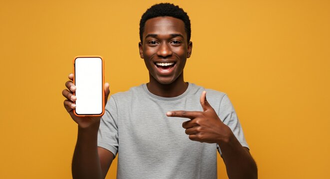 A cheerful young man in a gray t-shirt holds and points to a smartphone with a blank white screen against a vibrant yellow background, expressing excitement and enthusiasm.