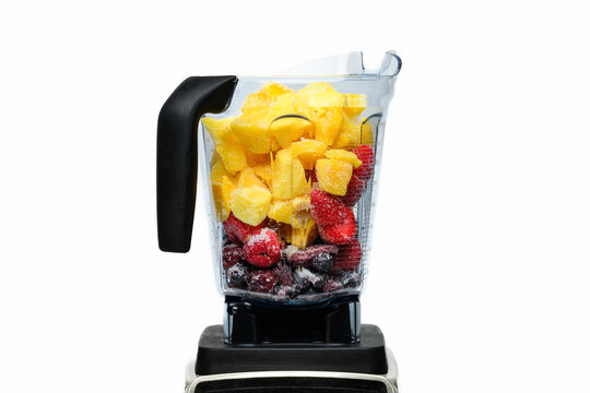 Frozen mangoes, blueberries, a&ccedil;ai fruit, and strawberries in a clear plastic blender against a white backdrop