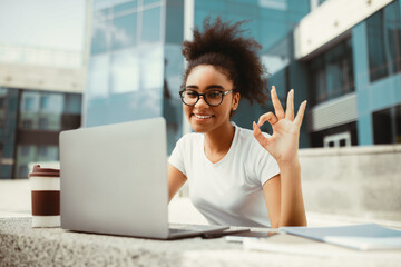 African American Female Student Gesturing Okay Using Laptop Approving E-Learning Sitting Outside Near Urban Univerity Building. Remote Learning And Modern Distance Education Concept