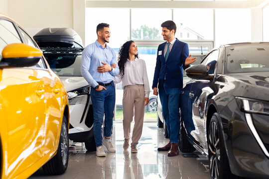Handsome arab guy sales assistant giding young family husband and wife by newest car salon, cheerful salesman showing appropriate black auto for happy middle-eastern couple, full length shot