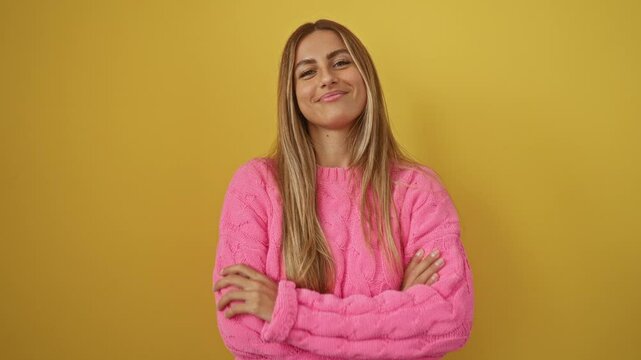 Woman, a beautiful young blonde person in sweater standing with crossed arms, smiling cheerfully and looking positive over isolated yellow background