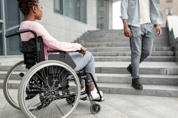 Obraz premium Young black disabled woman suffering from lack of wheelchair friendly facilities, looking at man going down stairs, cannot get home without ramp. Handicapped people problems concept