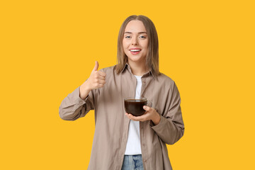 Beautiful young woman with glass cup of hot coffee showing thumb-up gesture on yellow background