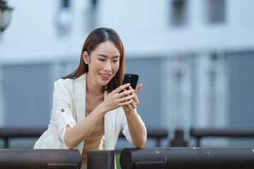 Fototapeta premium Asian businesswoman wearing a blazer is leaning on a railing and using her smartphone outdoors in an urban setting, smiling at what she's reading on the screen