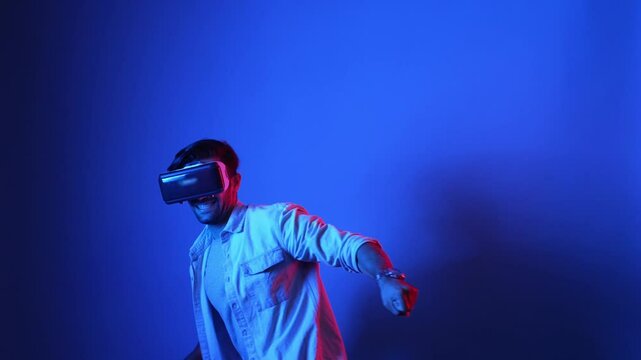 Smart gamer wearing VR goggles while holding sword and fencing in virtual world. Attractive male model playing action game while standing at blue background and accessing in metaverse. Apparatus.