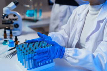 Scientist wearing blue gloves holds a rack of test tubes filled with blue liquid, conducting...