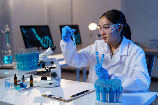 Female scientist wearing lab coat and gloves analyzing test tubes with blue liquid, conducting experiments and research in a modern laboratory with microscope and computer displaying dna structure
