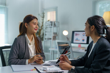 Two asian businesswomen are discussing work, using a tablet and a computer in a modern office, collaborating on a project and analyzing financial data