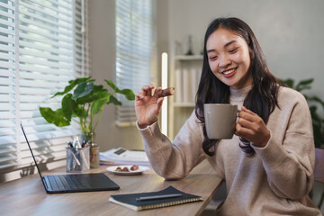 Young asian businesswoman enjoying a relaxing work break at home, savoring a chocolate bar and...