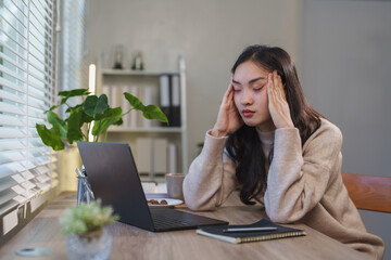Young asian businesswoman having a headache and massaging her temples while working on a laptop...