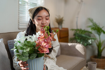 Young woman is sitting on her sofa in the living room, holding a metal vase with a bouquet of fresh flowers, enjoying their fragrance and smiling