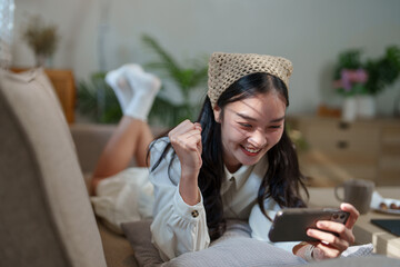 Happy young woman is lying on the sofa at home, raising her fist in celebration while playing...