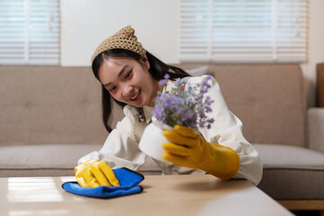 Asian housekeeper smiling and wearing yellow rubber gloves is cleaning a table and holding a flower...