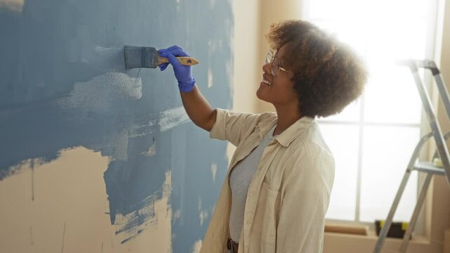 Woman painting wall indoors with blue paint in new home featuring curly hair wearing glasses and gloves in bright living room apartment setting with a ladder.