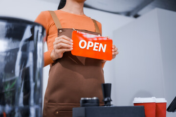 Asian barista with a beautiful face, wearing overalls, smiling warmly while holding a shop opening sign, welcoming customers to the counter bar at the front of a vibrant coffee shop