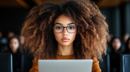 focused student with curly hair and glasses is sitting at a desk, actively using a laptop in a classroom filled with other students. atmosphere is serious and engaged