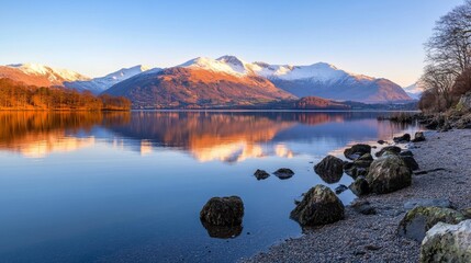 Sunrise over tranquil lake reflecting snow-capped mountains, calm waters, rocky shore, idyllic landscape