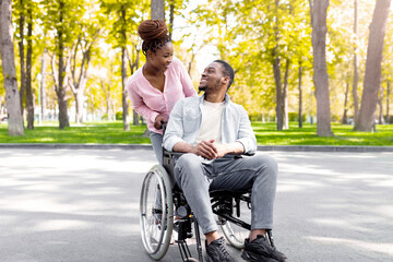 Full length of affectionate black woman and her disabled boyfriend in wheelchair looking at each other on walk at city park in autumn. Happy impaired couple enjoying fall nature outdoors