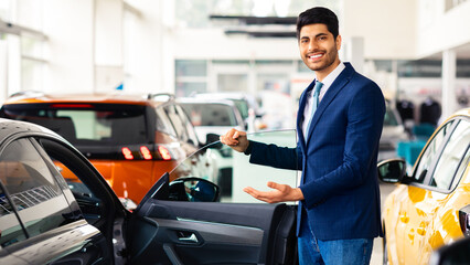 Cheerful young middle eastern man in formal outwear sales manager showing nice sports car, open black auto door and smiling, luxury automobile dealership salon interior, copy space