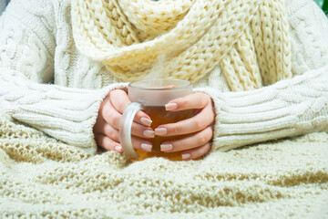 A woman in a large knit sweater holds a cup of hot tea in her hands. Hands close-up.
