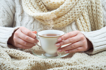 A woman in a large knit sweater holds a cup of hot tea in her hands. Hands close-up.