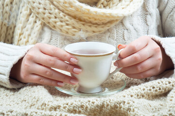 A woman in a large knit sweater holds a cup of hot tea in her hands. Hands close-up.
