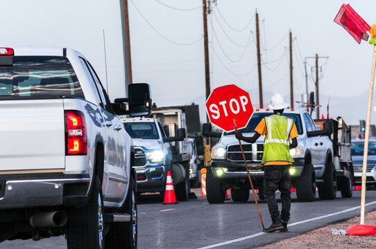 A construction worker in a reflective vest and hard hat holds a stop sign to direct traffic on a road under construction with utility poles and vehicles visible