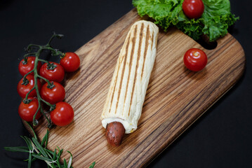 French hot dog with grilled sausage on a wooden board on a dark background, sauce, greens, tomatoes, vegetables, fast food