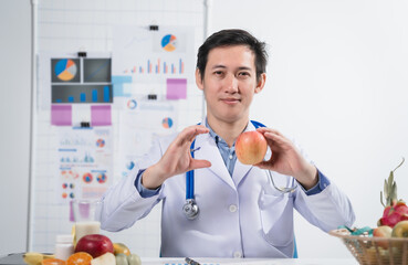 A male nutritionist working at a hospital desk, holding an apple and mixed fruit, offering care, health advice, weight loss guidance, and eating tips online for patients seeking nutrition support