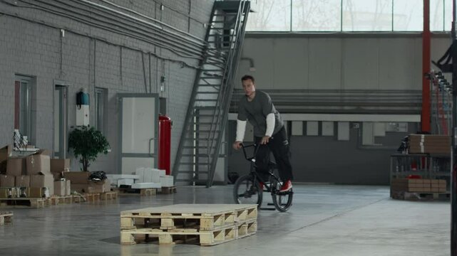 Full stab shot of young Caucasian guy stunt riding on BMX bicycle in hangar and failing to do clean spin, practicing with friend