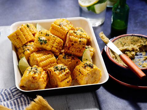 Freshly grilled corn on the cob is beautifully arranged on a serving platter, topped with herbs and garnished with lime wedges. A vibrant dipping sauce accompanies the dish