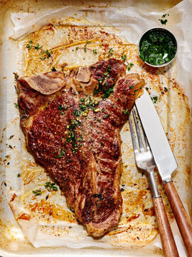 A perfectly cooked t-bone steak rests on a baking tray with herbs and spices, accompanied by a small bowl of green chimichurri sauce, ready to be enjoyed