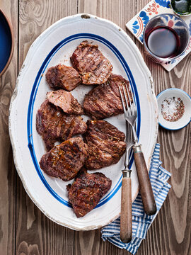 Succulent pieces of grilled meat rest on a rustic white platter accompanied by wine and garnishes. The warm wooden table creates a inviting atmosphere for a meal