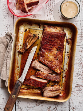 Tender, smoked ribs are sliced and displayed on a metal tray, surrounded by barbecue sauce and a refreshing drink, perfect for a summer backyard feast