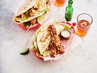 Two baskets filled with grilled chicken skewers and tacos sit on a table. Lime wedges complement the meal, along with cold beverages in glasses and a bottle