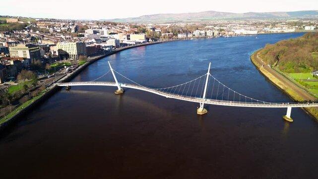Wide retreating aerial shot of the Peace Bridge in Derry-Londonderry, NI on a bright and sunny day. Filmed in 4K at 30fps and in Rec709 color space.