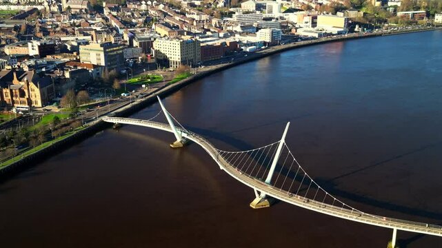 Aerial shot of the Peace Bridge in Derry-Londonderry, NI. Shot in a wide, the camera shows the bridge and city on a bright sunny day. Filmed in 4K at 30fps and in Rec709 color space.