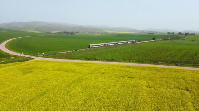 The scene is an aerial view of a bridge crossing over a valley with a train track running through it. On the other side of the valley, there are green fields and mountains in the distance.