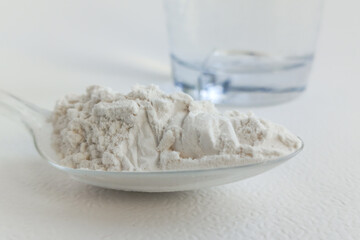 A plastic spoon with protein powder lies on a white table, with a glass of water in the background. Dry protein powder. Gain of muscle mass. A food additive.