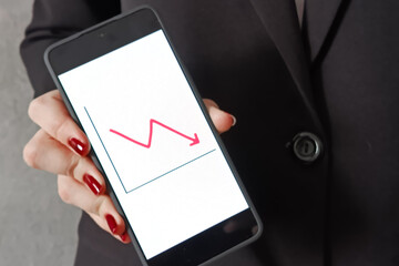 A smartphone with a graph falling down in a woman's hand on the screen, with a woman in a business suit in the background. The concept of income reduction, failure. Financing. 
