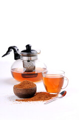 a glass teapot filled with amber-colored tea, a glass cup of tea, a bowl of brown sugar, and a spoon,