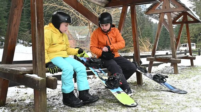 Two figures, one adult and one child, sit on a wooden bench in snowy surroundings. They are dressed in winter sports gear and appear to be preparing their snowboards.