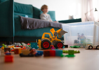 A toy tractor and building blocks scattered on the floor in a living room, with a child sitting on the couch in the background. Concept of everyday life, children's mess, and family routine.