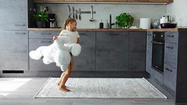 Little girl dancing with big white teddy bear in the home kitchen in the morning
