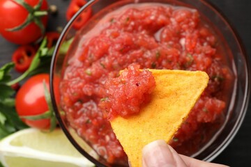 Woman dipping nacho chip into spicy salsa sauce at table, top view