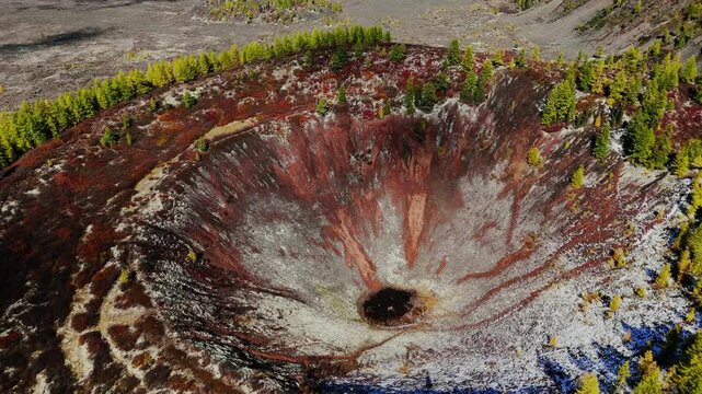 Aerial view of a large volcanic crater with vivid shades of rusty red, gray, and black. The crater is surrounded by an autumn forest with golden and green trees, 