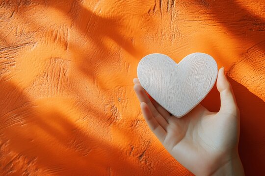 Hand holding white heart shaped object against textured orange wall with soft light - Powered by Adobe