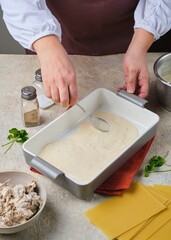 Step by step preparation of lasagna with mushrooms and chicken on a light concrete background. Putting béchamel sauce on the bottom of a ceramic baking dish. Step by step recipes.