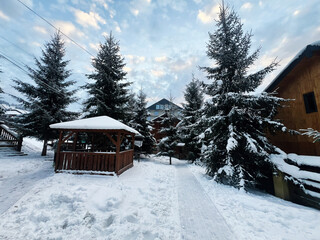 Snow-covered gazebo with evergreen trees and houses in a peaceful winter setting