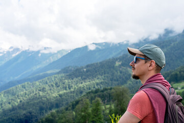 traveler with backpack stands breathtaking mountain landscape Wearing green cap , red shirt, man looks over green hills , peaks under cloudy sky. outdoor adventure, beauty of nature copy space